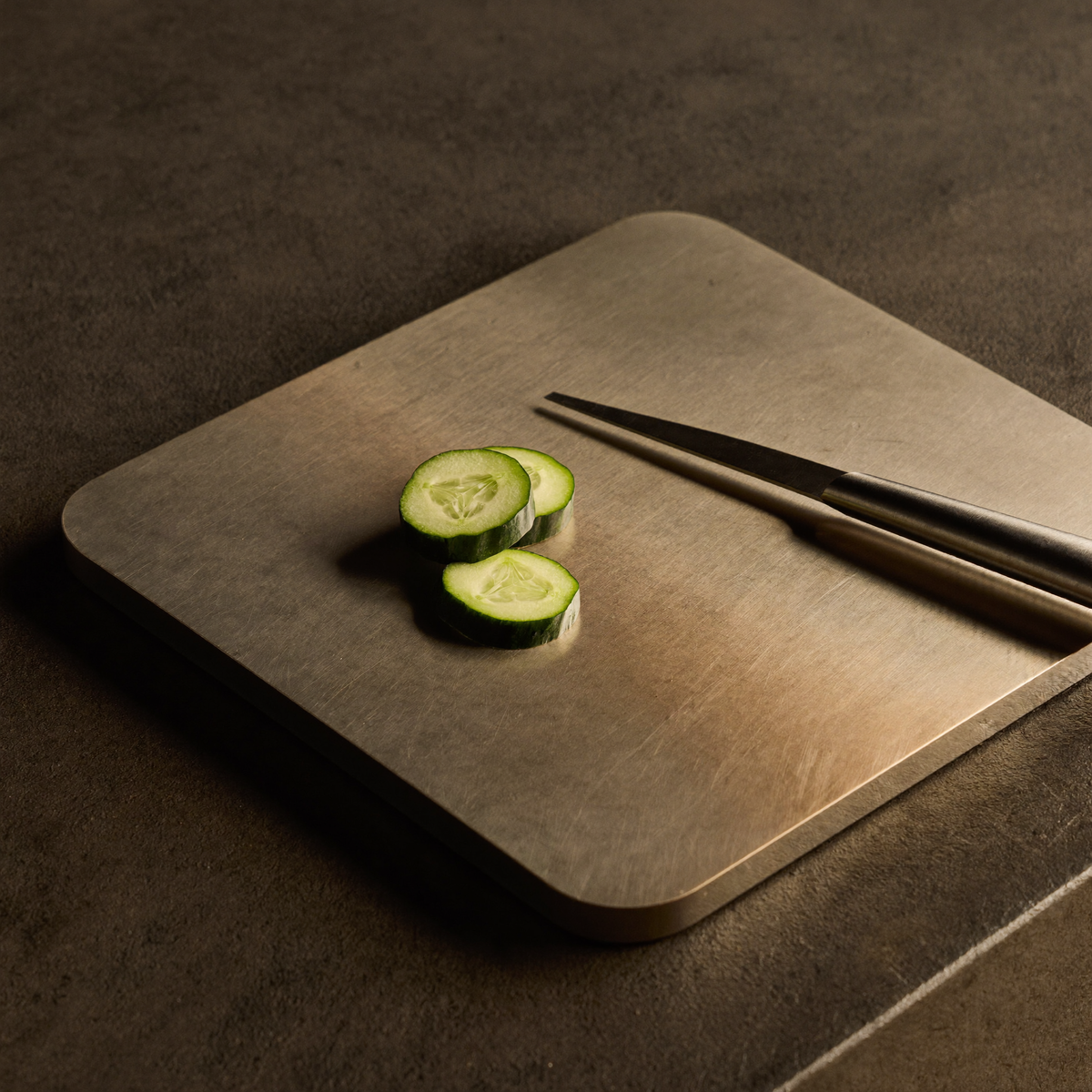 Planche à découper en titane brossé sur un plan sombre, avec des rondelles de concombre et un couteau